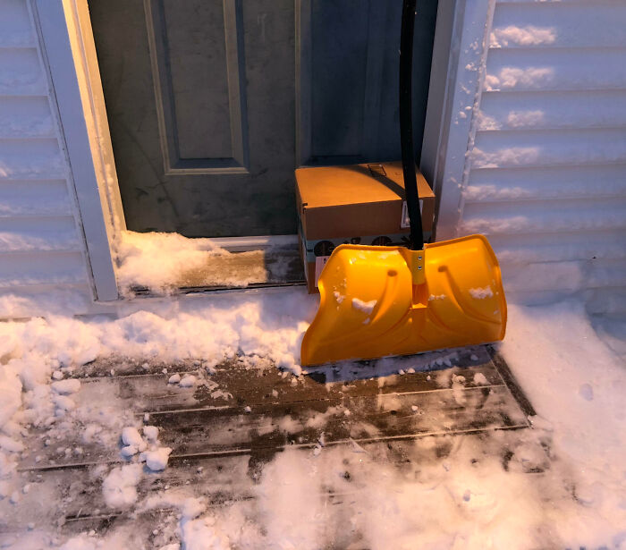 Yellow snow shovel leaning on a doorstep with snow around and a delivery box, showing things that happen only in Canada.