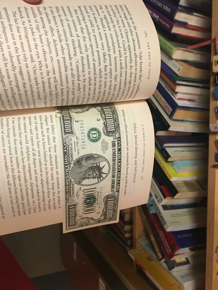 A book with a cool bookmark resembling a $100,000 bill, surrounded by a shelf of other books.