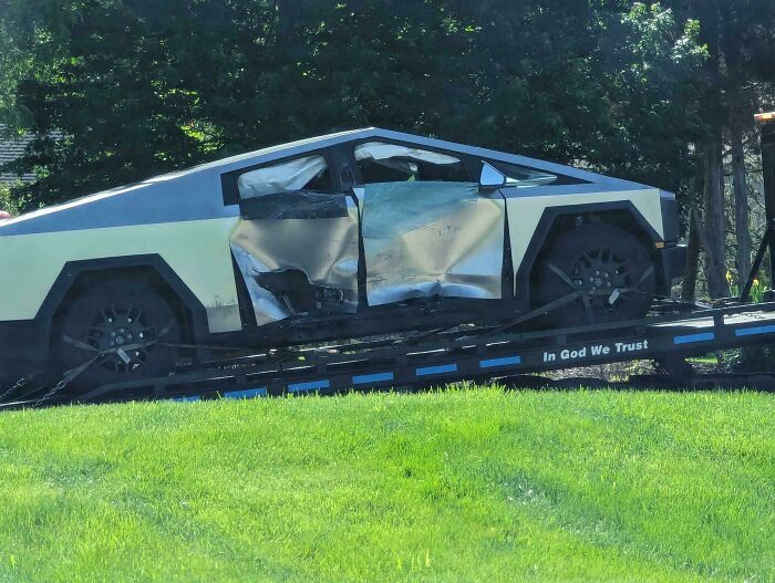 Damaged futuristic car on a towing trailer, highlighting an expensive accident amid a green landscape.