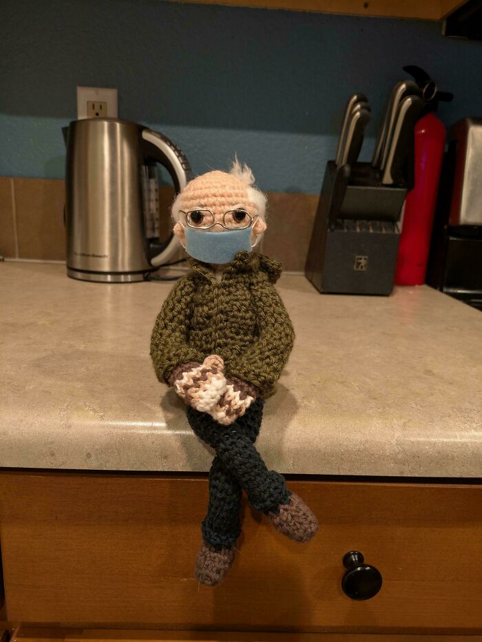 Crochet figure resembling a man with glasses and mask, wearing green sweater, sitting on a kitchen countertop.