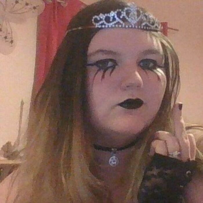 Teen girl with dark gothic makeup and tiara showing middle finger in a regrettable past pic that evokes cringe and humor.