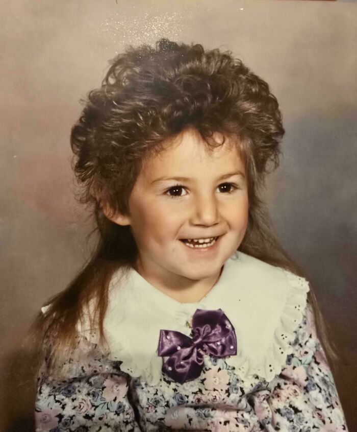 Child with a regretful past wearing a floral dress and purple bow, smiling with a voluminous 80s hairstyle.