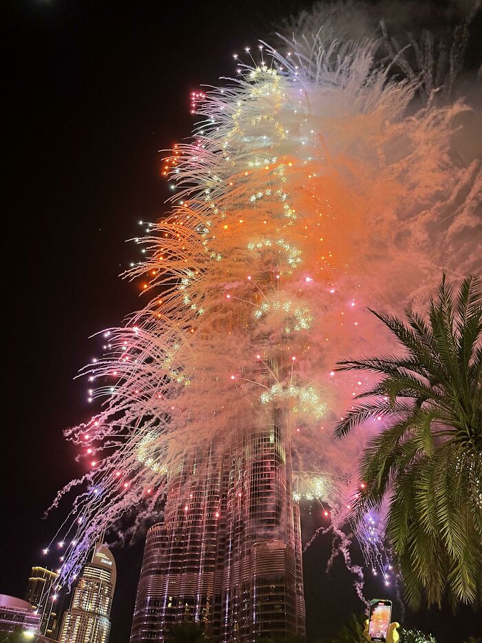 Fireworks display around the Burj Khalifa in Dubai at night, with colorful lights and palm tree in the foreground.