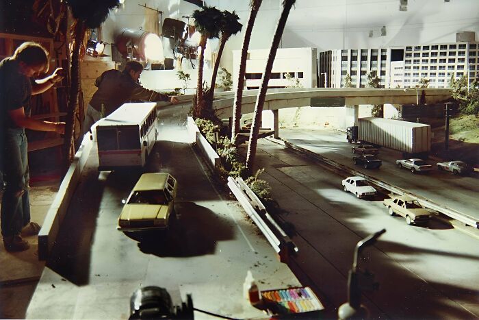 Two men adjusting a detailed 90s miniature cityscape model with cars and palm trees in an indoor setting.
