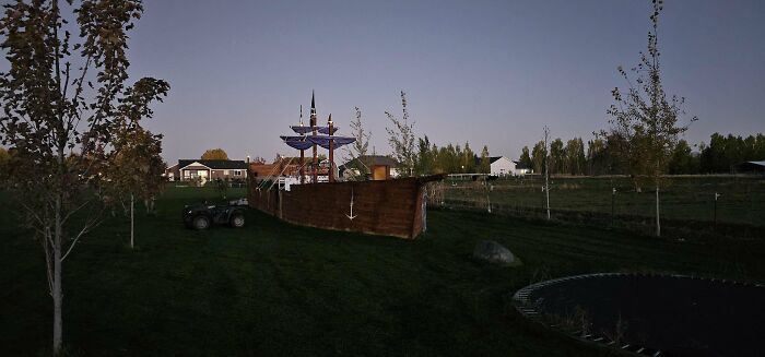 A backyard with a large wooden playground ship, surrounded by trees and grass, showcasing a dad's rewarding creativity.