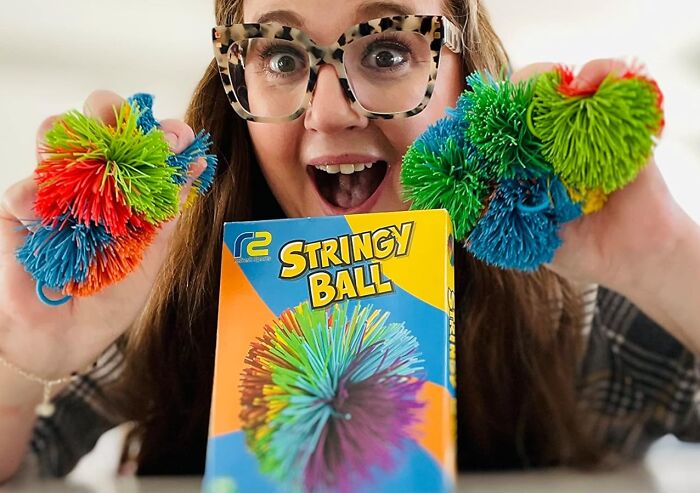 Woman excitedly playing with colorful 90s Stringy Ball toys, smiling with glasses.