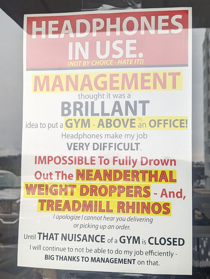 Sign with funny passive-aggressive message about office noise from gym above, mentioning headphones and management decisions.