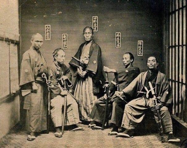 Samurai in traditional attire with swords posed inside a wooden room, showcasing a historical perspective.