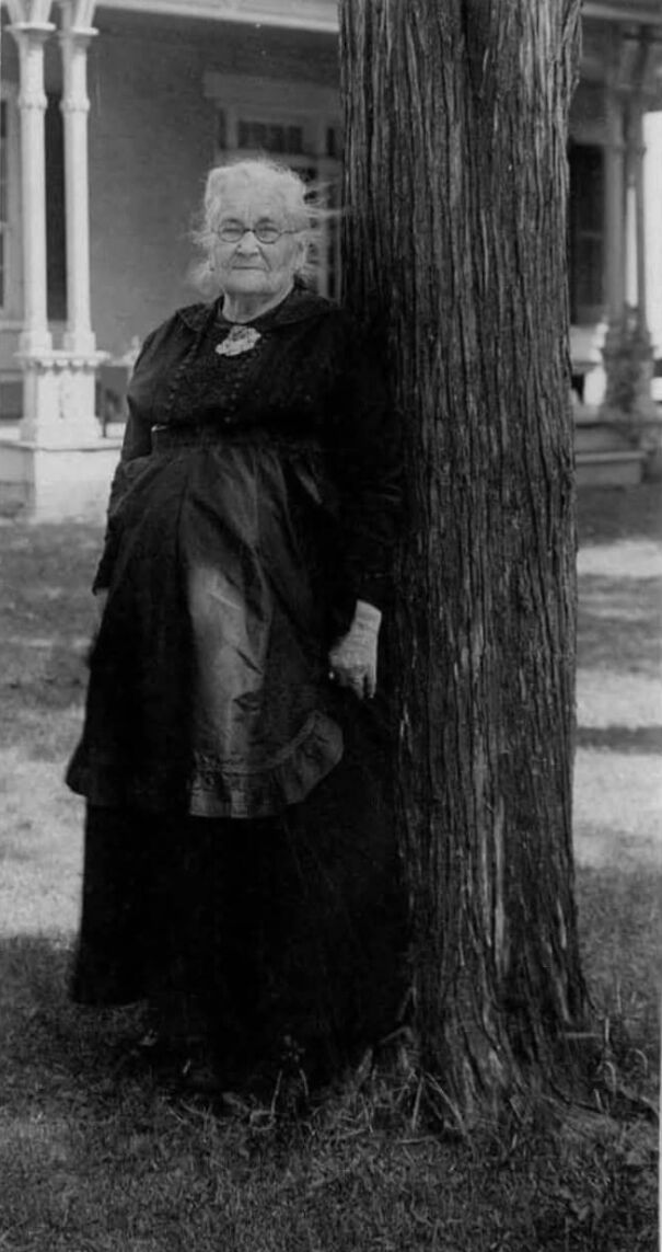 Elderly woman in vintage attire leaning against a tree, representing lesser-known historical moments.