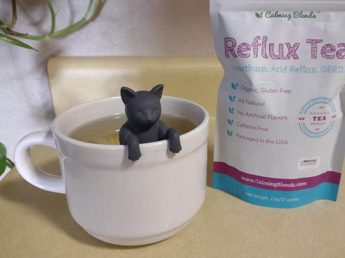 Genuine Fred cat-shaped tea infuser in a cup beside a Reflux Tea package on a kitchen counter.
