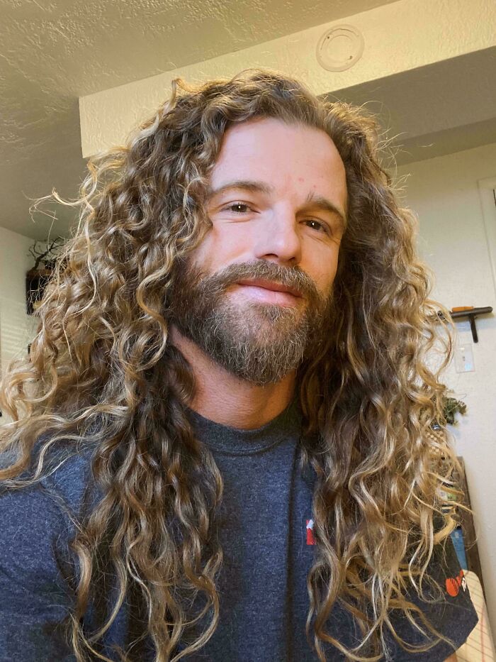 Man with long curly hair and beard smiling indoors, showcasing fierceflow beautiful hair men style and natural texture.