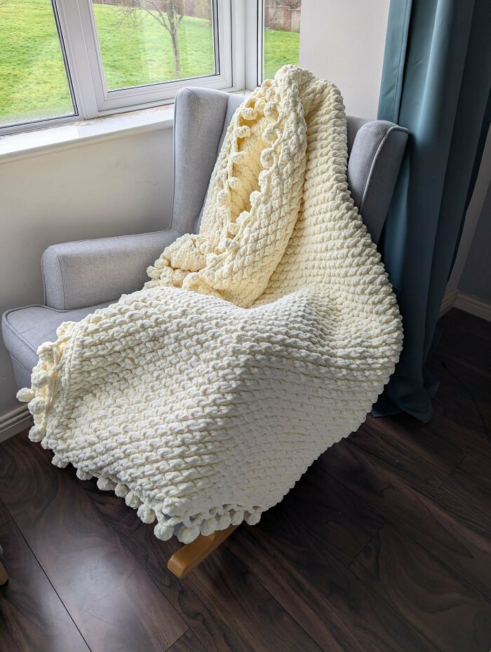 Chunky cream crochet blanket with textured pattern draped over a gray armchair by a window, showcasing crochet craftsmanship.