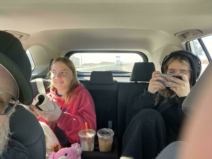 Dad capturing parenting moments with two kids in the backseat, one reading and one wearing headphones playing on a phone.