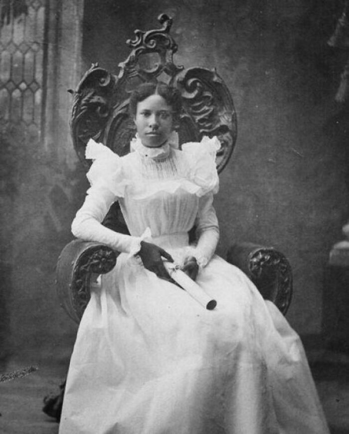 Young woman in a vintage white dress sitting in an ornate chair, holding a rolled document in candid glimpses into the past.
