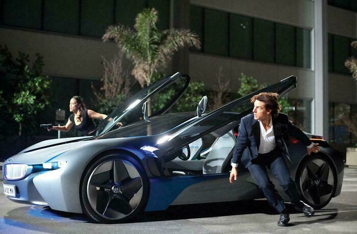 Actors escaping a futuristic car with gullwing doors in a scene highlighting questionable movie details mocked by cinephiles online.