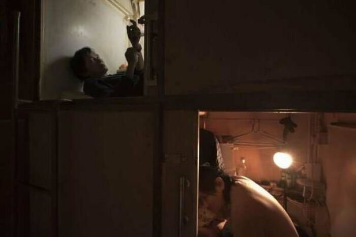 Two individuals in cramped urban living spaces, one lying above and the other sitting below with a light. Urban hell depiction.
