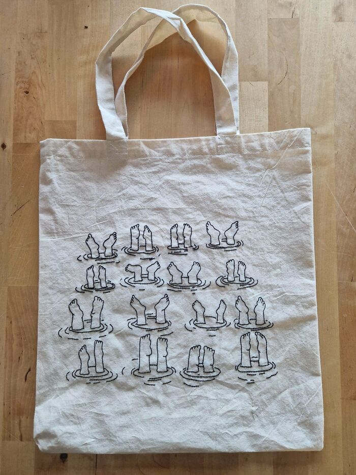 Beige tote bag featuring embroidery of various small feet and legs poking out of water in black thread.