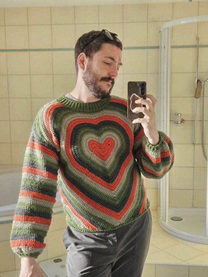 Man taking a selfie wearing a colorful heart-patterned crochet sweater, showcasing impressive crochet craftsmanship.