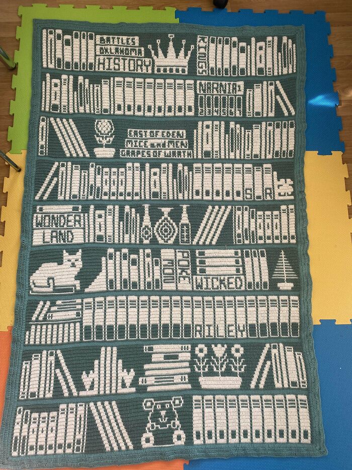 Crochet blanket designed to look like bookshelves filled with books, flowers, and decorative items, showcasing intricate crochet skills.