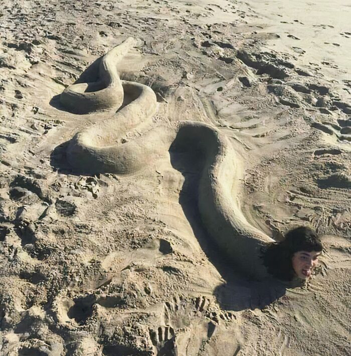 Persona con cabeza emergiendo de un cuerpo de arena en la playa, creando una ilusión impactante.