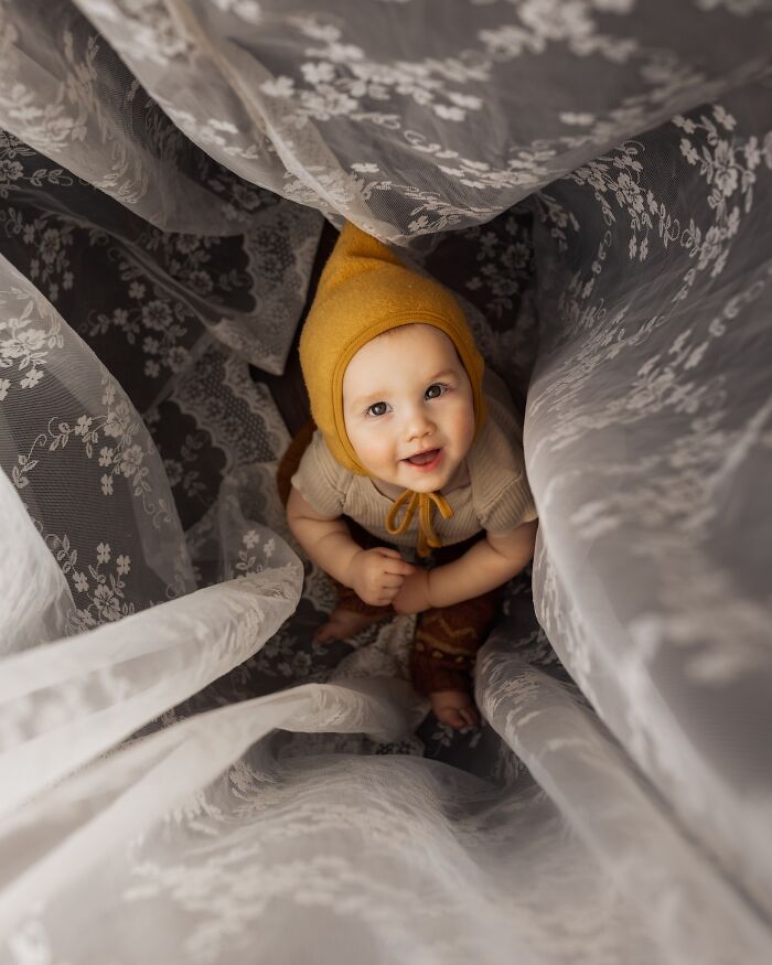 Childhood magic captured as a baby in a yellow bonnet peeks through lace curtains, smiling up warmly.