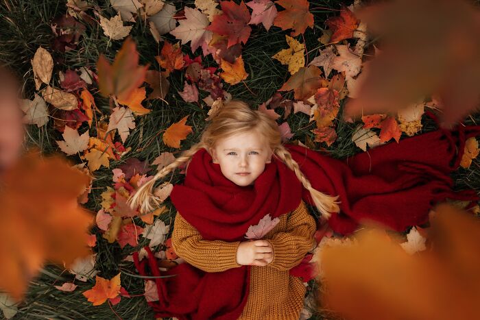 Child in a red scarf lies on autumn leaves, capturing the magic of childhood through a mother's lens.