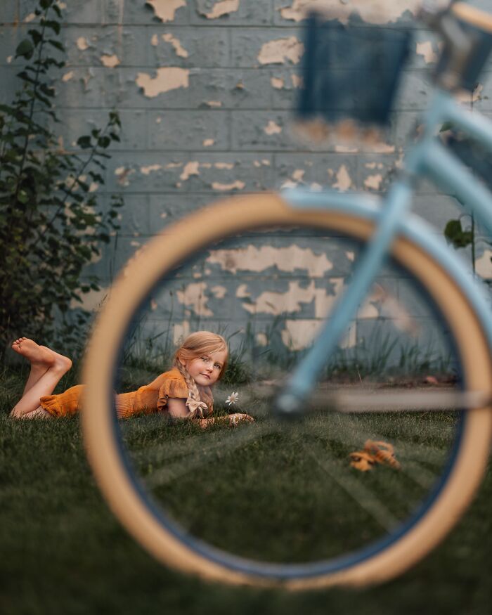 Childhood magic captured as a girl in a yellow dress lies on grass, framed by a blue bike wheel.