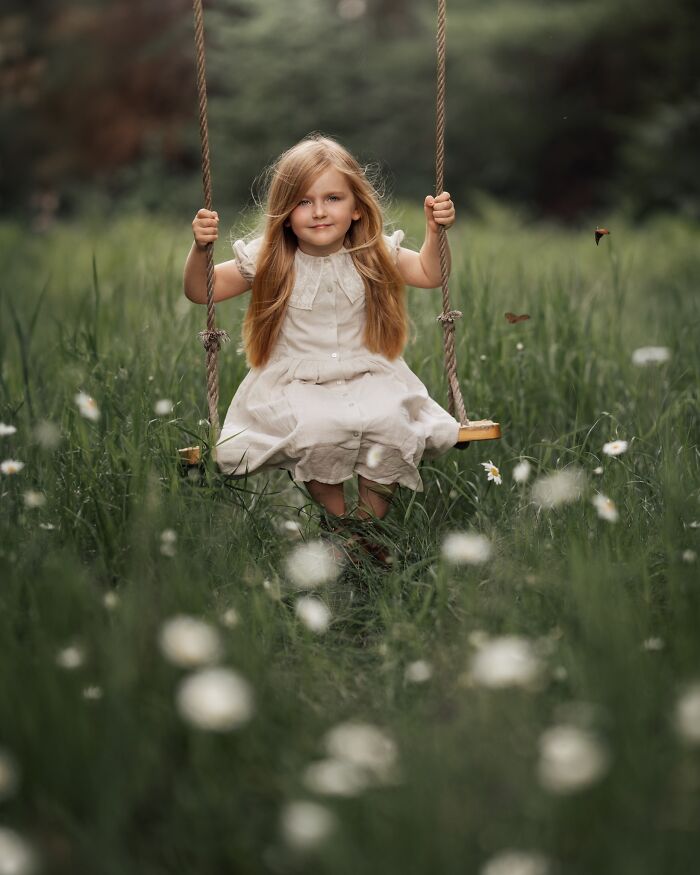 Child in a white dress on a swing, surrounded by grass and flowers, portraying childhood magic.