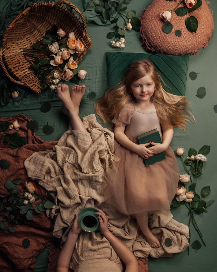 A serene childhood moment captured, with a young girl in a soft dress holding a book amidst flowers and fabrics.