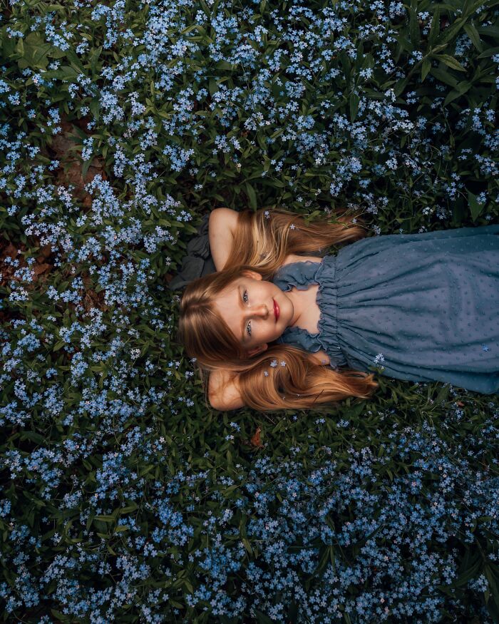 Young girl lying in a field of blue flowers, capturing the magic of childhood through a mother’s lens.