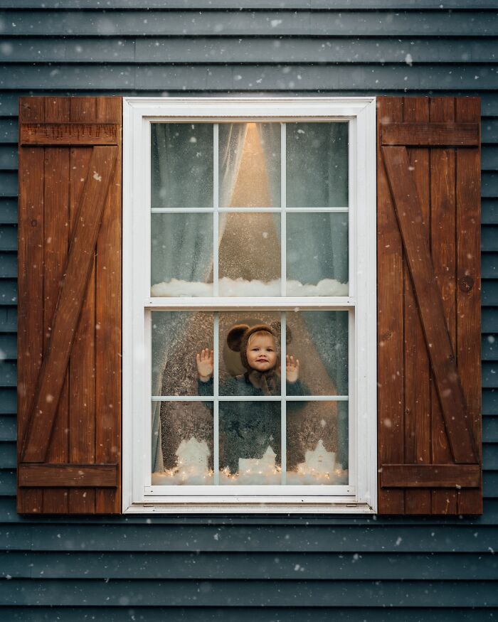Child in bear costume at window, touching glass with snow falling outside, capturing magic of childhood.