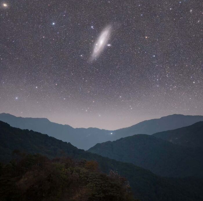 Astrophotography of a starry night sky with a visible galaxy above silhouetted mountain ridges.