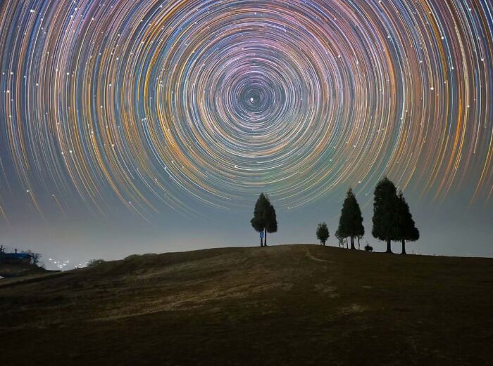 Astrophotography featuring vivid star trails swirling above silhouetted trees on a hill.