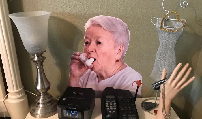 Cardboard cutout of a woman using an inhaler placed on a cluttered nightstand with lamps and clocks.