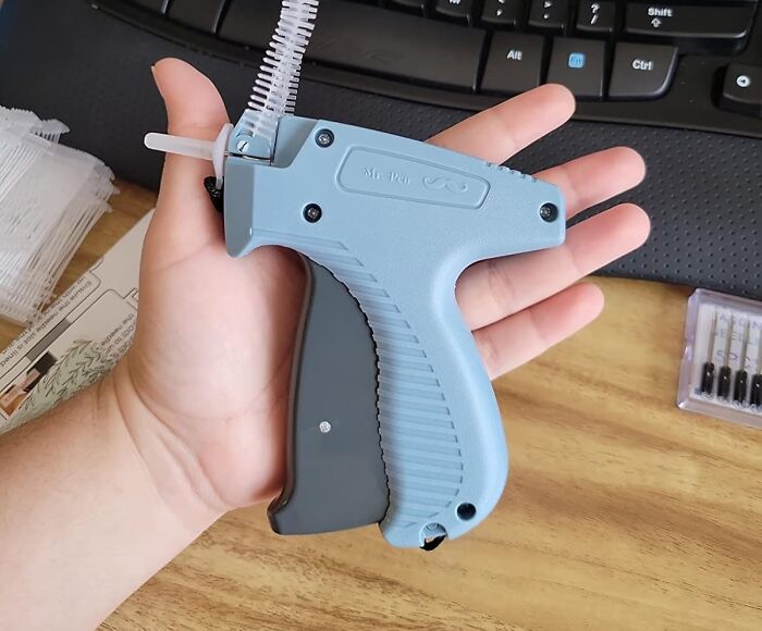 Hand holding a blue tagging gun, influenced by TikTok trends, next to a keyboard on a desk.