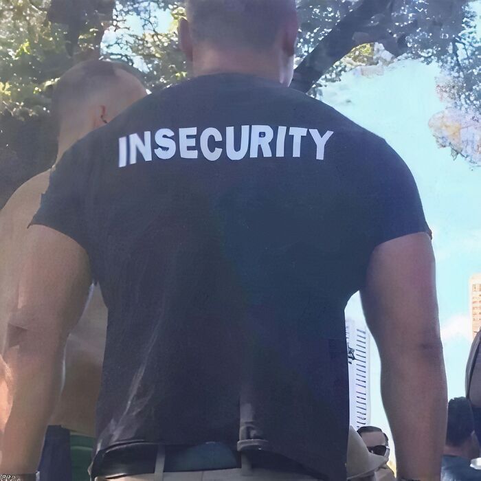 Man wearing a chaotic T-shirt with "Insecurity" printed on the back, standing outdoors under trees.