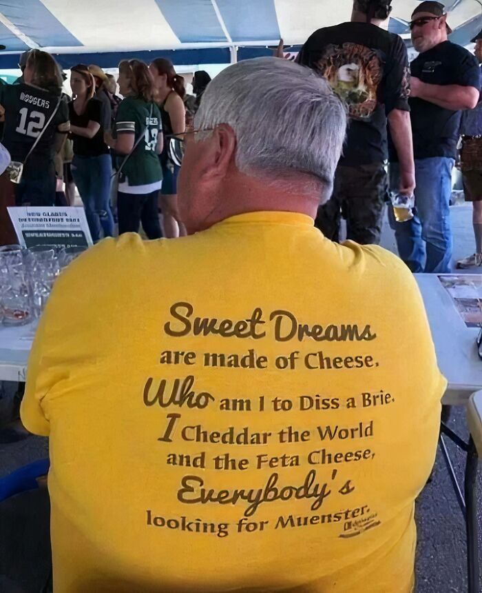 Man wearing a chaotic yellow T-shirt with cheese-themed parody text at an outdoor gathering.