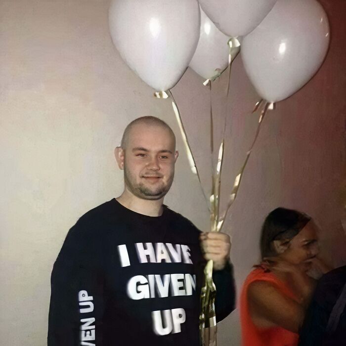 Man holding balloons, wearing a chaotic T-shirt with "I Have Given Up" printed on it, standing in front of a plain wall.