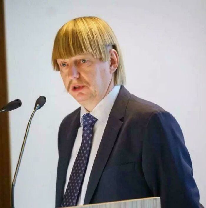 A man with an unfortunate bowl haircut and blonde bangs, speaking at a podium.