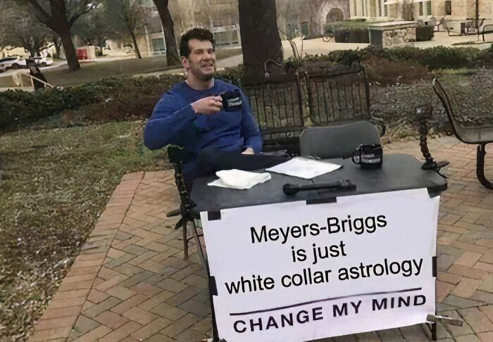 Man seated outdoors with a "Meyers-Briggs is just white collar astrology" sign, enjoying coffee, illustrating work-related humor.