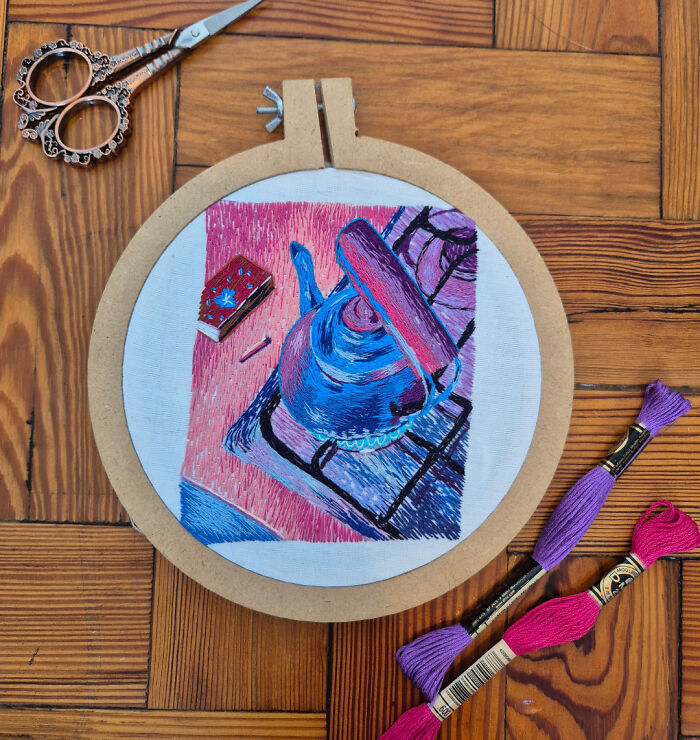 Embroidery hoop with a colorful stitched kettle design, surrounded by scissors and embroidery threads on a wooden floor.