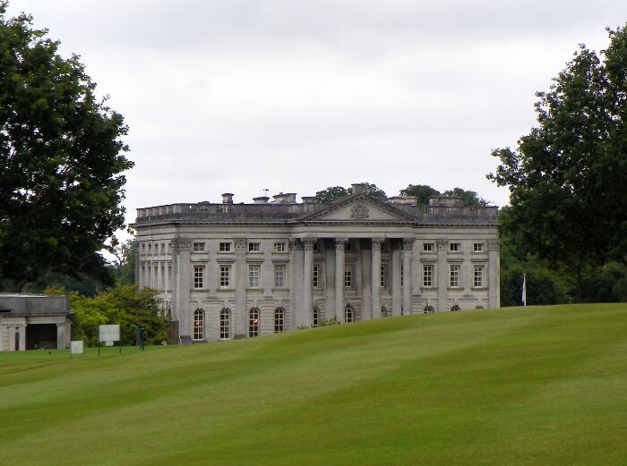 Historic mansion overlooking lush green golf course, surrounded by trees and cloudy sky.