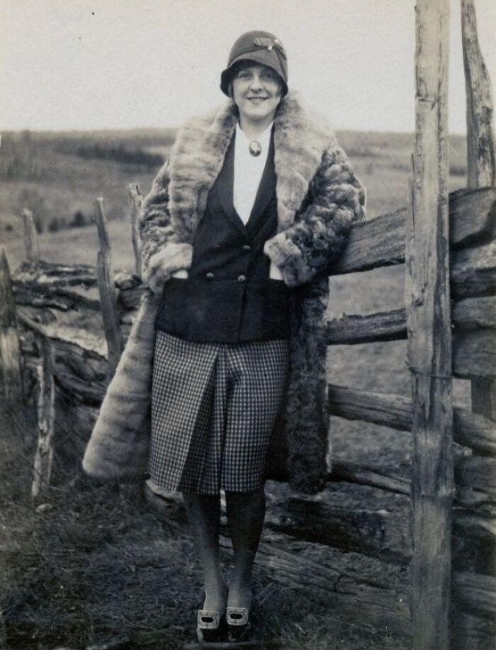 A woman in vintage fashion with a fur coat and hat, standing by a wooden fence from 50-100 years ago.