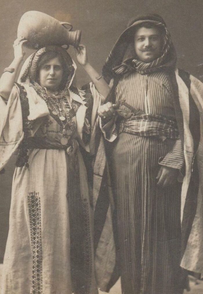 Two people in traditional attire, woman holding a jug on head, embodying rare historical fashion from decades past.