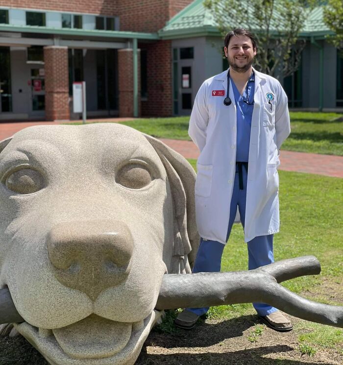 Veterinarian in scrubs and white coat smiling beside a large dog statue outside the office.