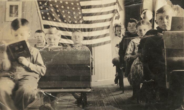 Children in an old classroom sitting at wooden desks with an American flag in the background, showcasing historical change.
