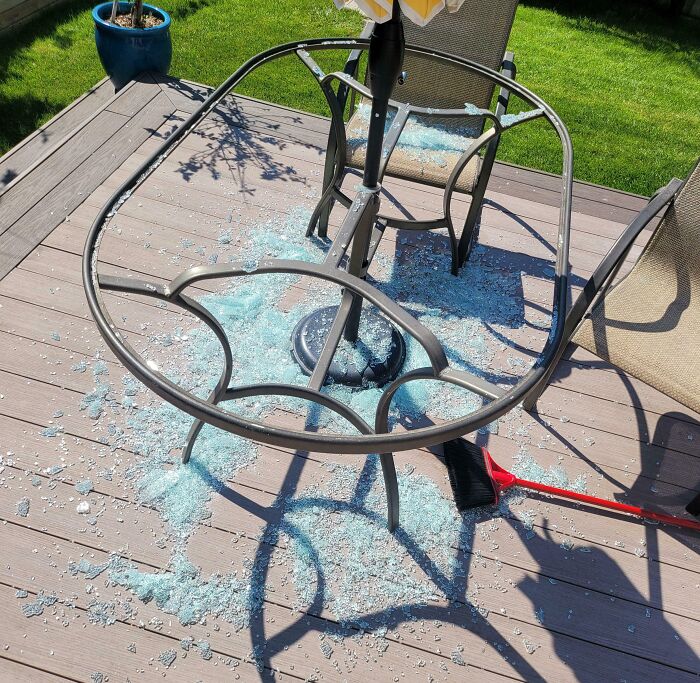 Shattered glass table on a wooden deck, remnants of an expensive accident, with a red broom nearby.