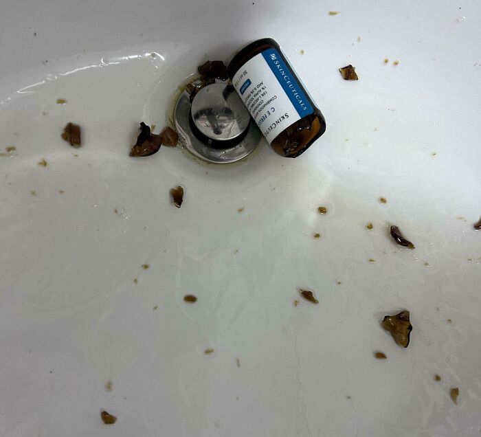 Broken glass bottle in sink, expensive accident with skincare product scattered around the drain.