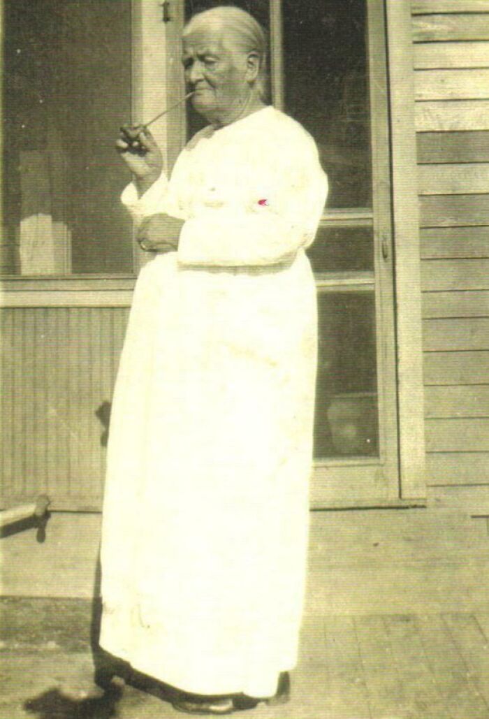 Elderly person from rare historical photo, standing in a long white gown, smoking a pipe outside a wooden house.