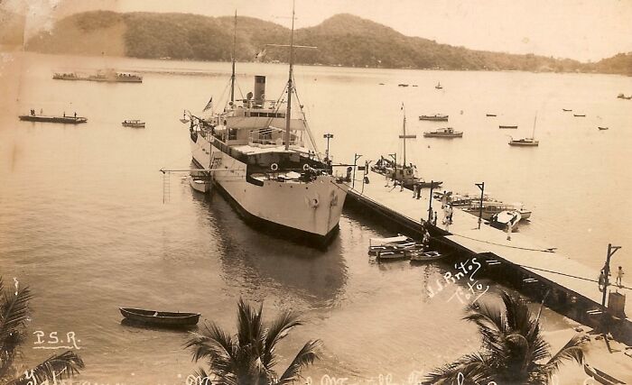 Vintage photo of a large ship docked in a harbor with small boats around, showcasing how the world has changed.